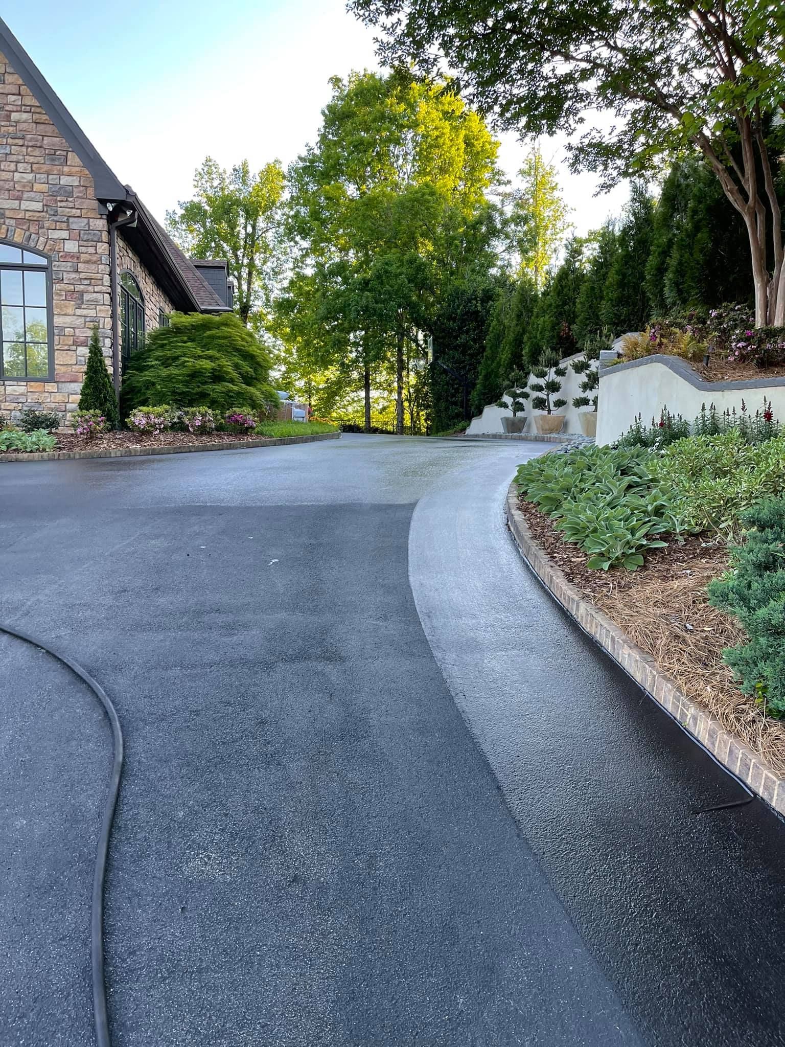 A stone house exterior features a curved asphalt driveway bordered by a landscaped retaining wall and lush green trees.