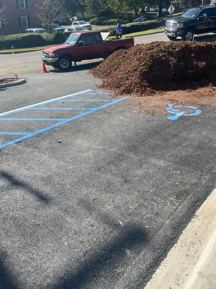 A large pile of mulch blocks a blue-painted accessible parking space in a parking lot next to a parked red pickup truck. A large pile of mulch blocks a blue-painted accessible parking space in a parking lot next to a parked red pickup truck.
