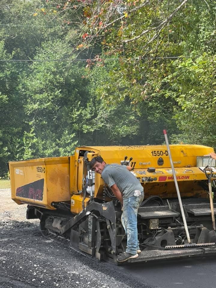A person operates a yellow asphalt paver on a road construction site surrounded by trees. A person in work clothes stands on a yellow construction paver on a paved road surrounded by trees.