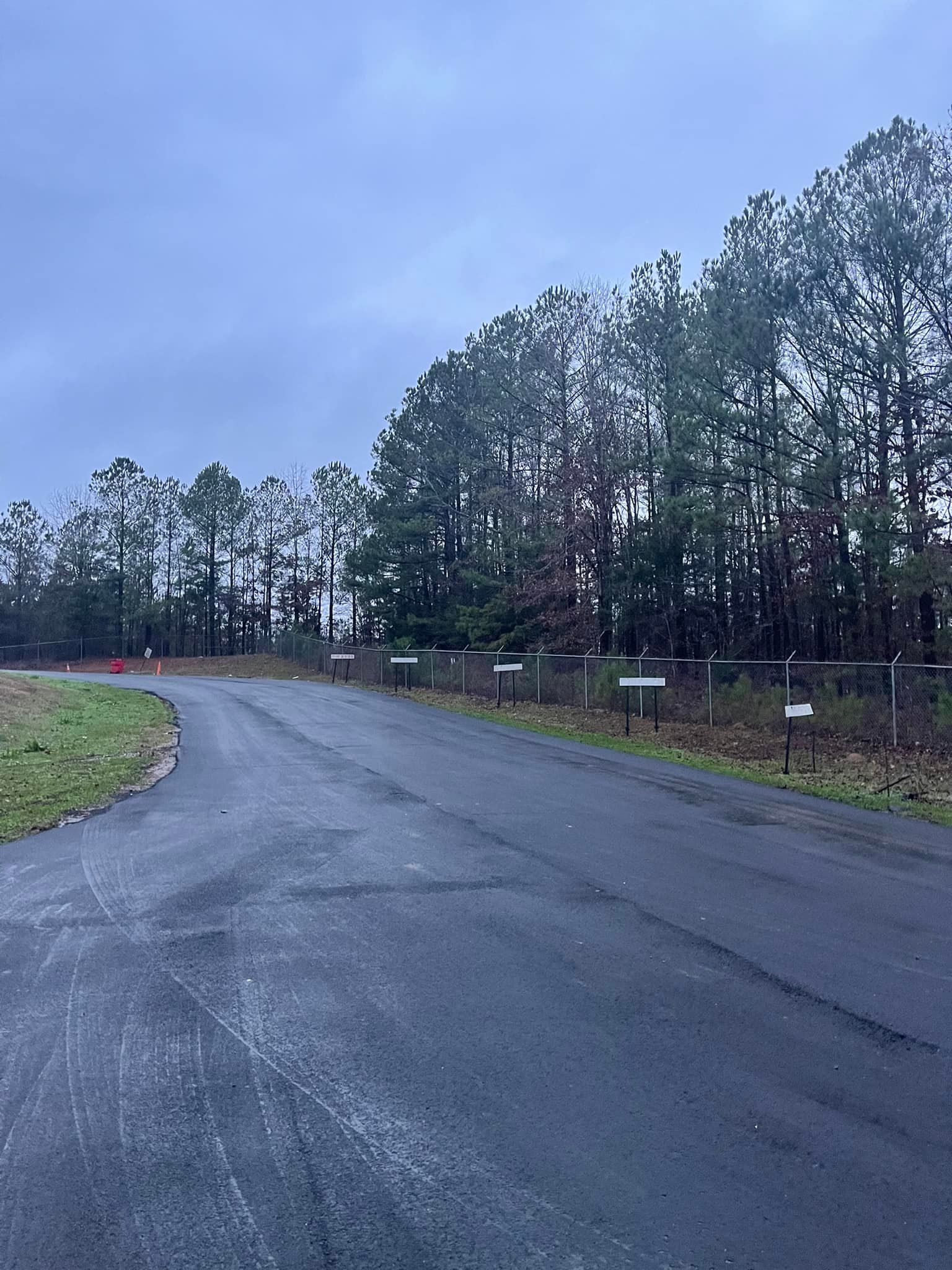 An asphalt road curves past a line of trees with small, evenly spaced signs along the edge on an overcast day.