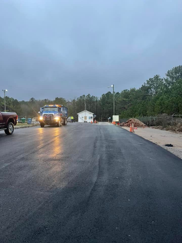 A dump truck drives on a freshly paved black road towards a small building at dusk under a cloudy sky.