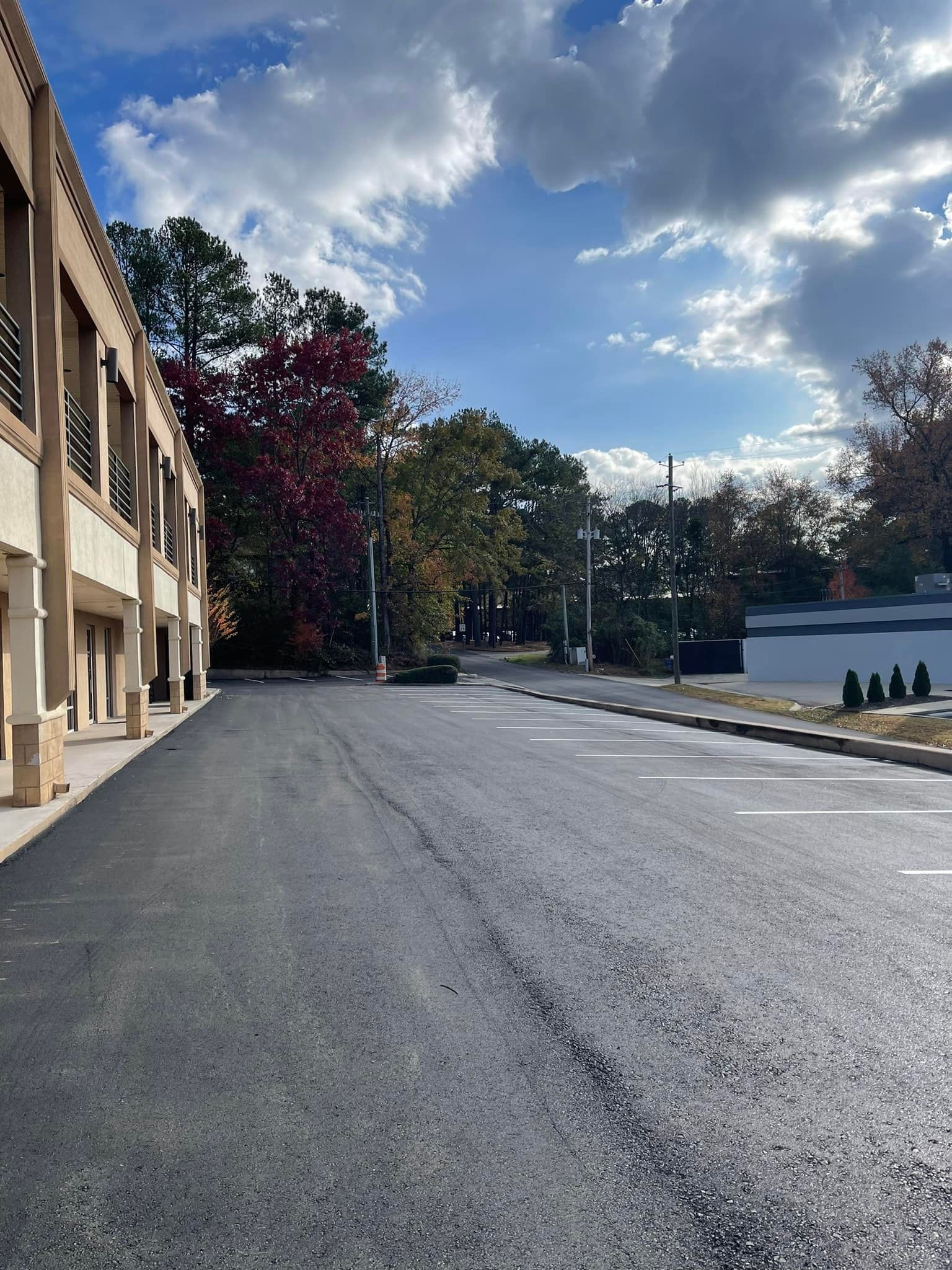 A freshly paved asphalt parking lot next to a modern building with arched columns under a bright, partly cloudy sky.