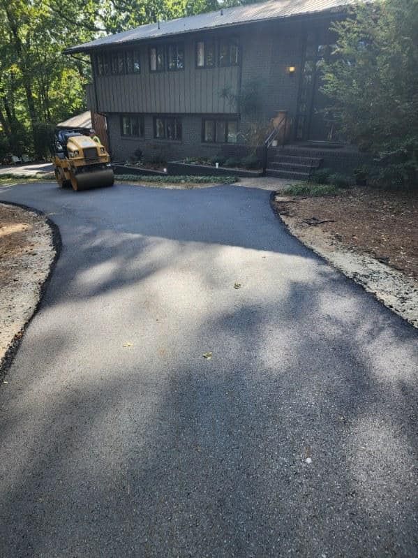 A yellow construction roller paving a fresh asphalt driveway leading to a multi-story home surrounded by trees.