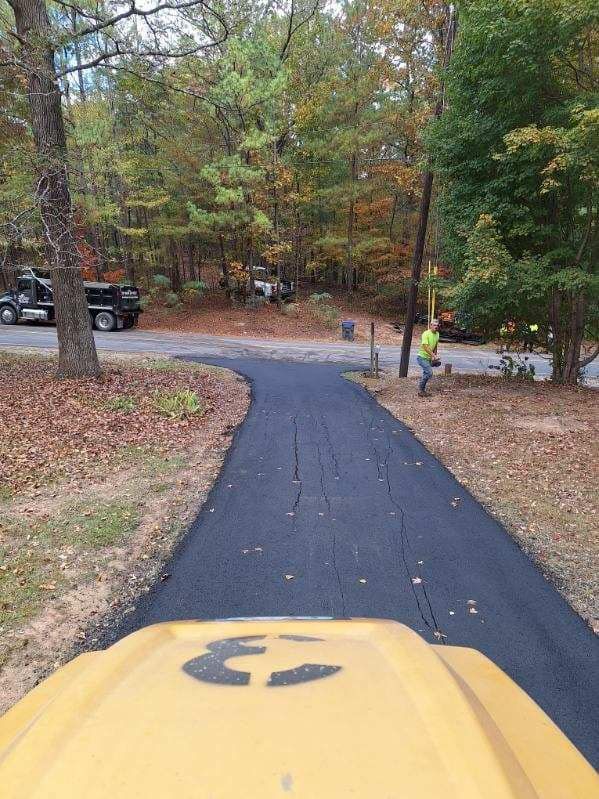 A view from a yellow construction vehicle looking down a freshly paved asphalt driveway toward a tree-lined road.