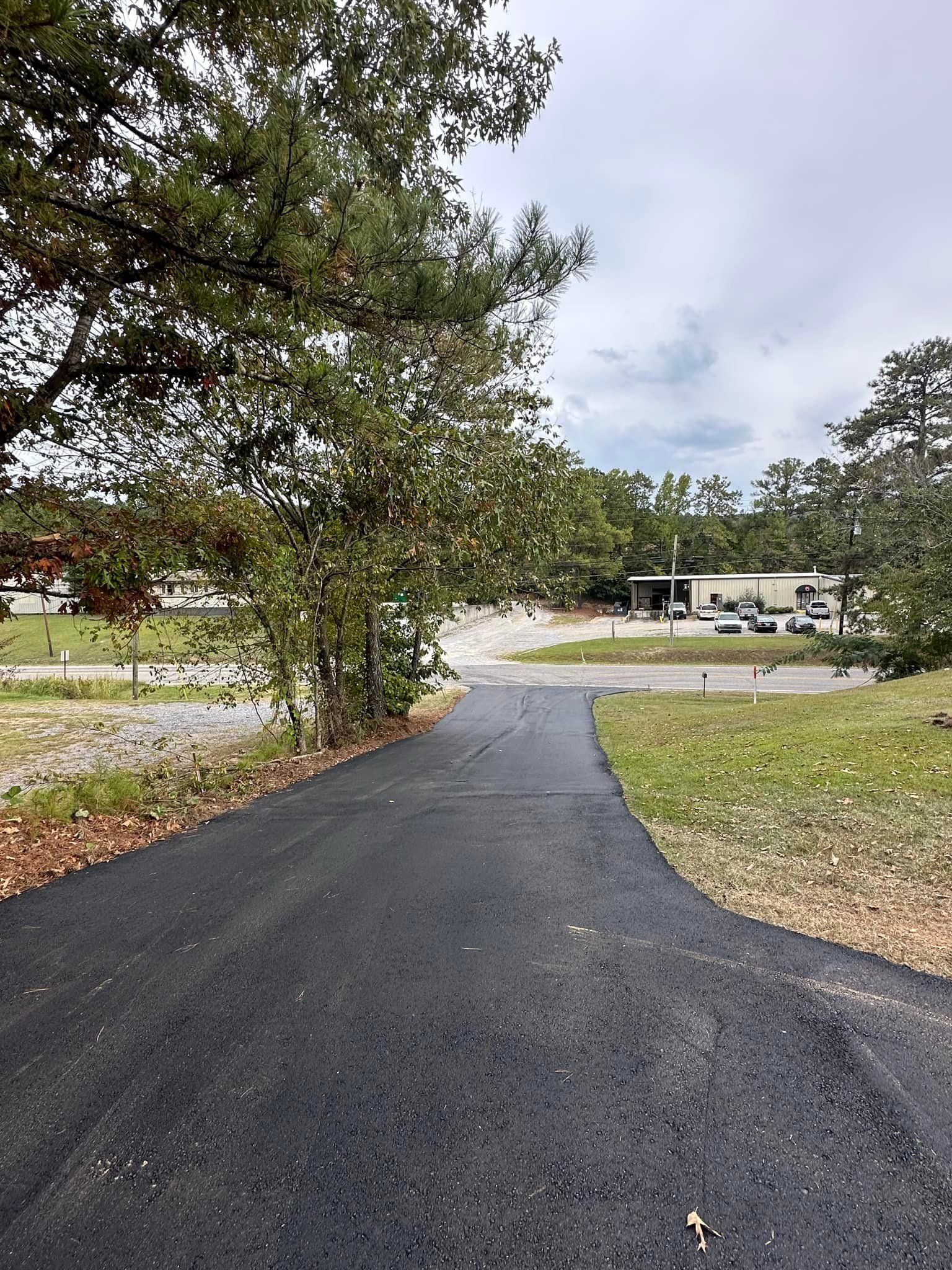 A newly paved black asphalt driveway leads toward a gravel road and buildings under a cloudy sky.