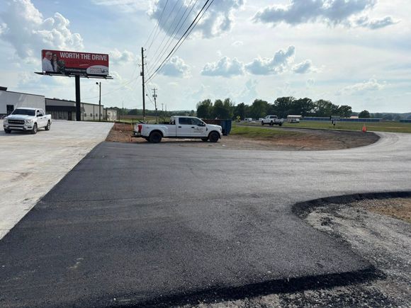 A freshly paved asphalt driveway leads to a parking area with white pickup trucks under a partly cloudy sky. A freshly paved asphalt driveway leads to a parking area with white pickup trucks under a partly cloudy sky.