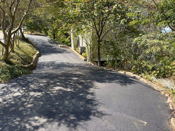 A freshly paved black asphalt driveway winds upward through a sunlit, tree-lined residential area. A freshly paved black asphalt driveway winds upward through a sunlit, tree-lined residential area.