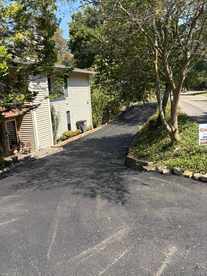 A paved driveway slopes downhill toward a light-colored two-story house, bordered by trees and stone landscaping.
