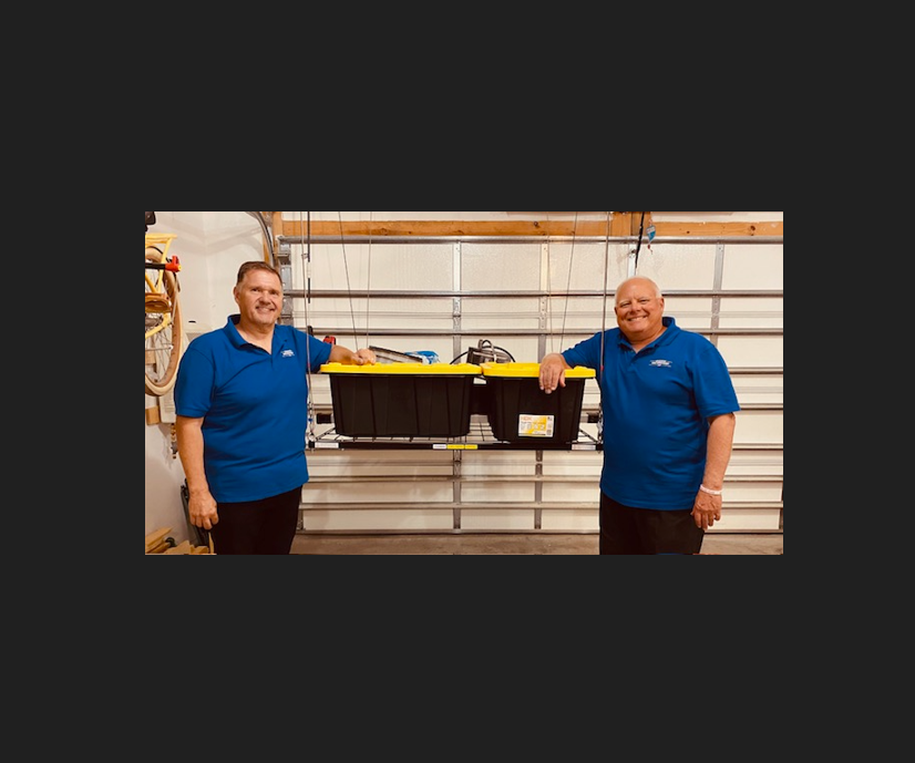 Two men in blue shirts holding a black storage bin in a garage