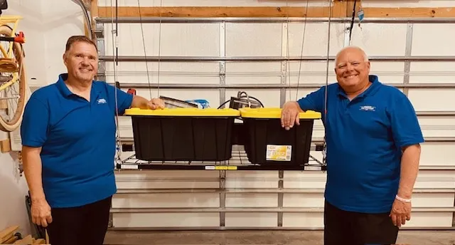 Two men in blue shirts holding a black bin in a warehouse aisle