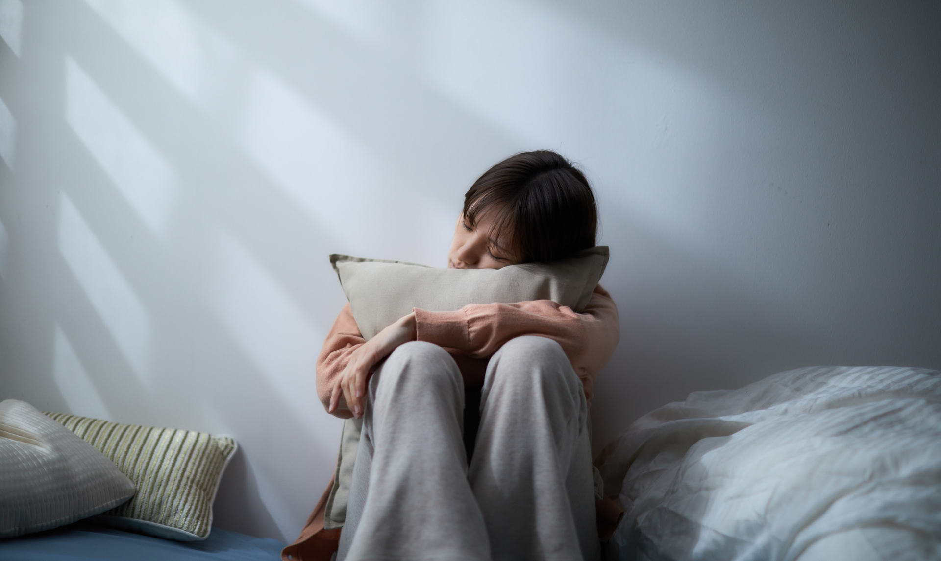 Person seated, hugging pillow, near a wall. Harsh light, shadow stripes. Appears distressed. Person seated, hugging pillow, near a wall. Harsh light, shadow stripes. Appears distressed.