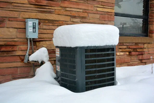 Un climatiseur est recouvert de neige sur le côté d'un bâtiment en briques.