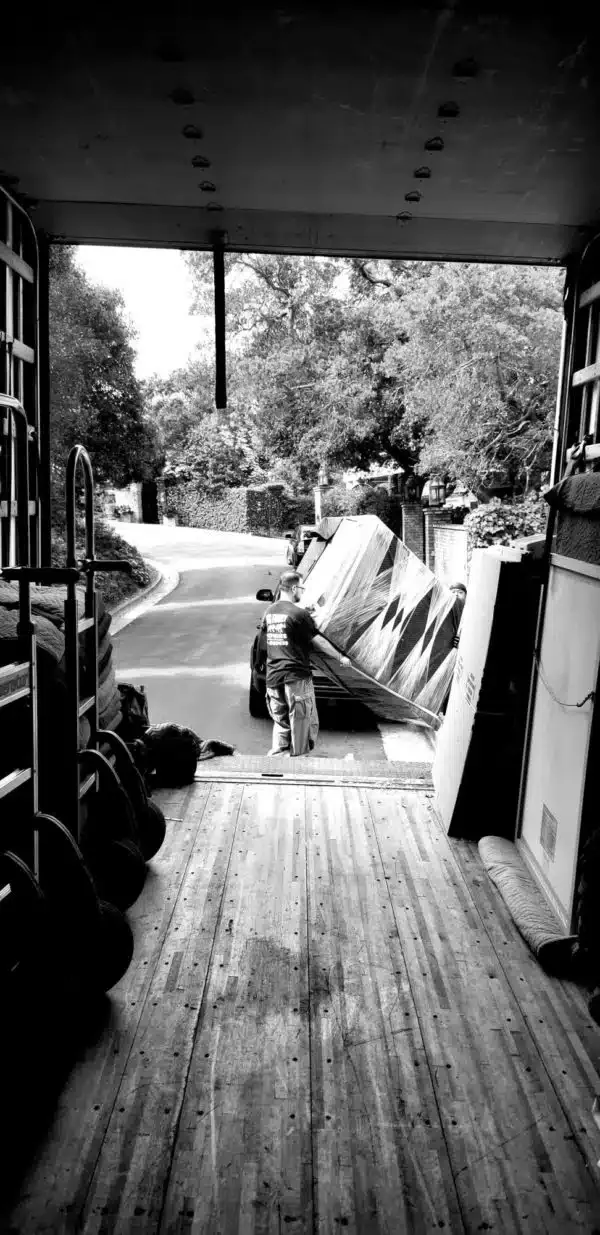 Covered porch with wooden floor, chairs, and stacked boards overlooking a tree-lined driveway.