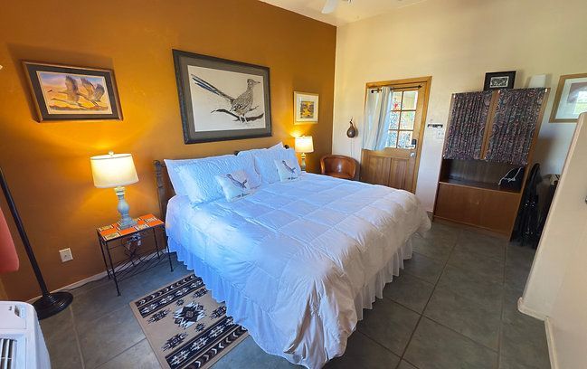 Warm-toned bedroom with a made bed, bedside lamp, framed wall art, and a dresser near a window