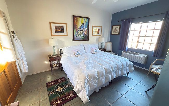 Bright bedroom with a neatly made white bed, colorful wall art, and a window seat by a sunny window.