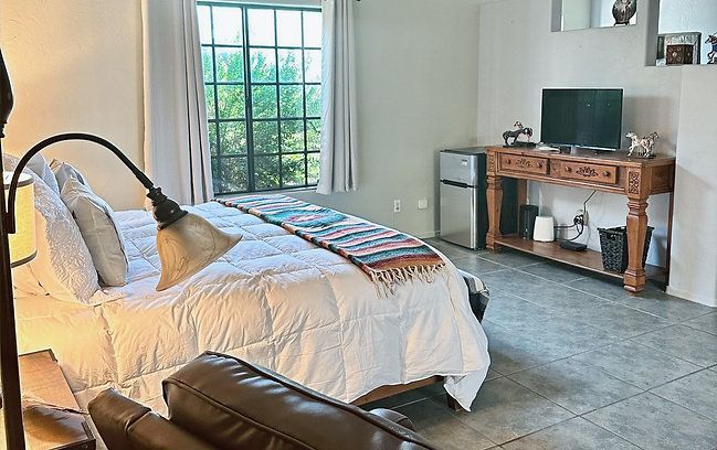Bright bedroom with a white bed, floor lamp, window, leather chair, and wooden TV stand on tiled floor.