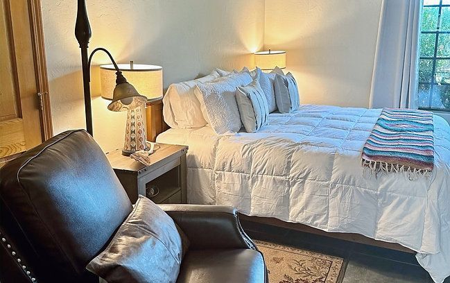 Cozy bedroom with a white bed, bedside lamp, dark leather chair, and folded throw by a window.