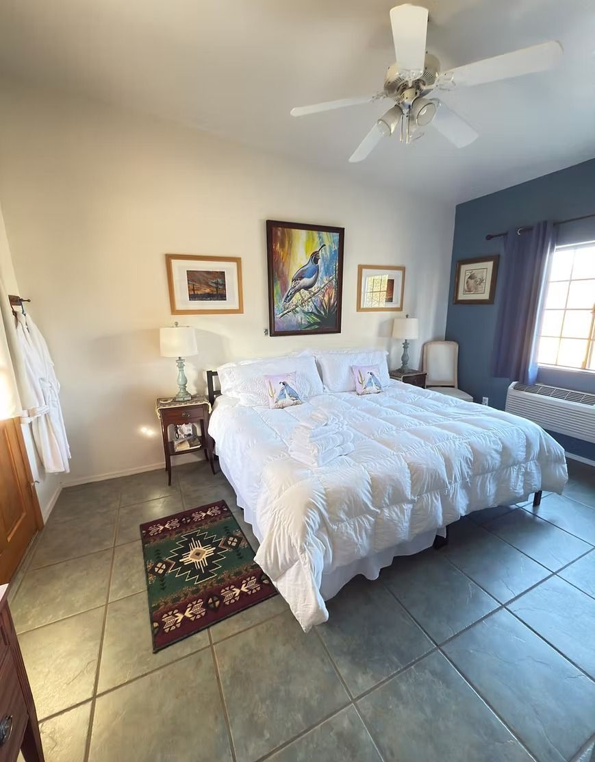 Bright bedroom with a white bed, tiled floor, ceiling fan, and windows with blue curtains.
