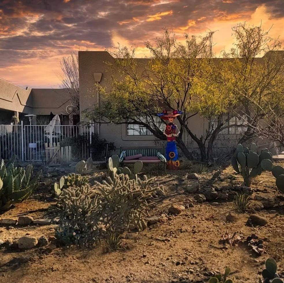 Desert front yard at sunset with a small colorful sculpture, cactus, and a house in the background