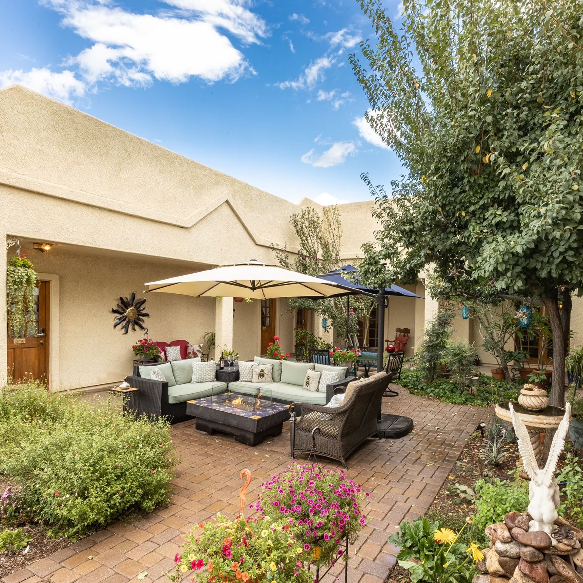 Outdoor patio with wicker seating, umbrellas, flowers, and a large tree beside a stucco house