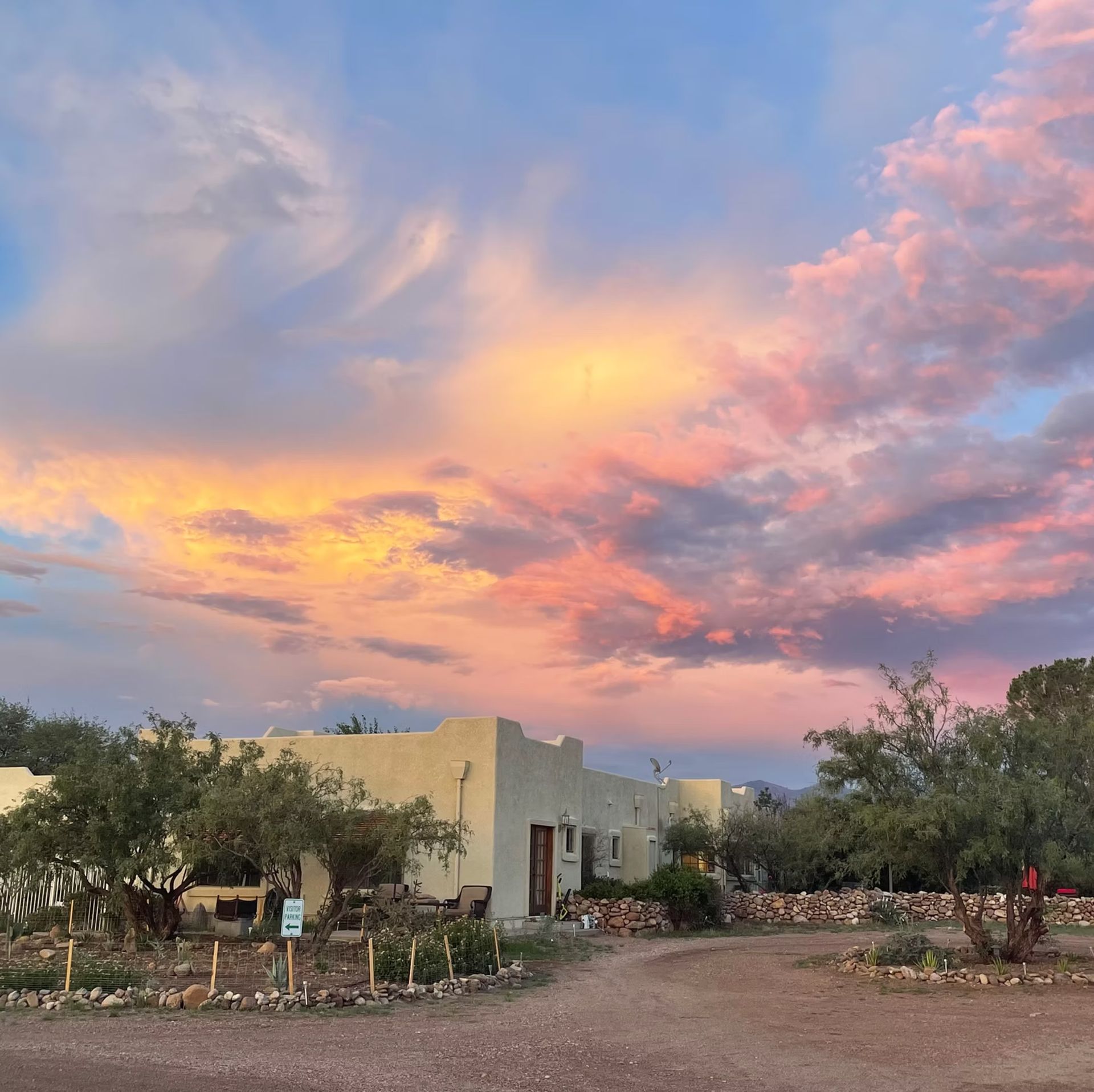 Desert home at sunset under a pastel pink and orange sky, with trees and a dirt driveway
