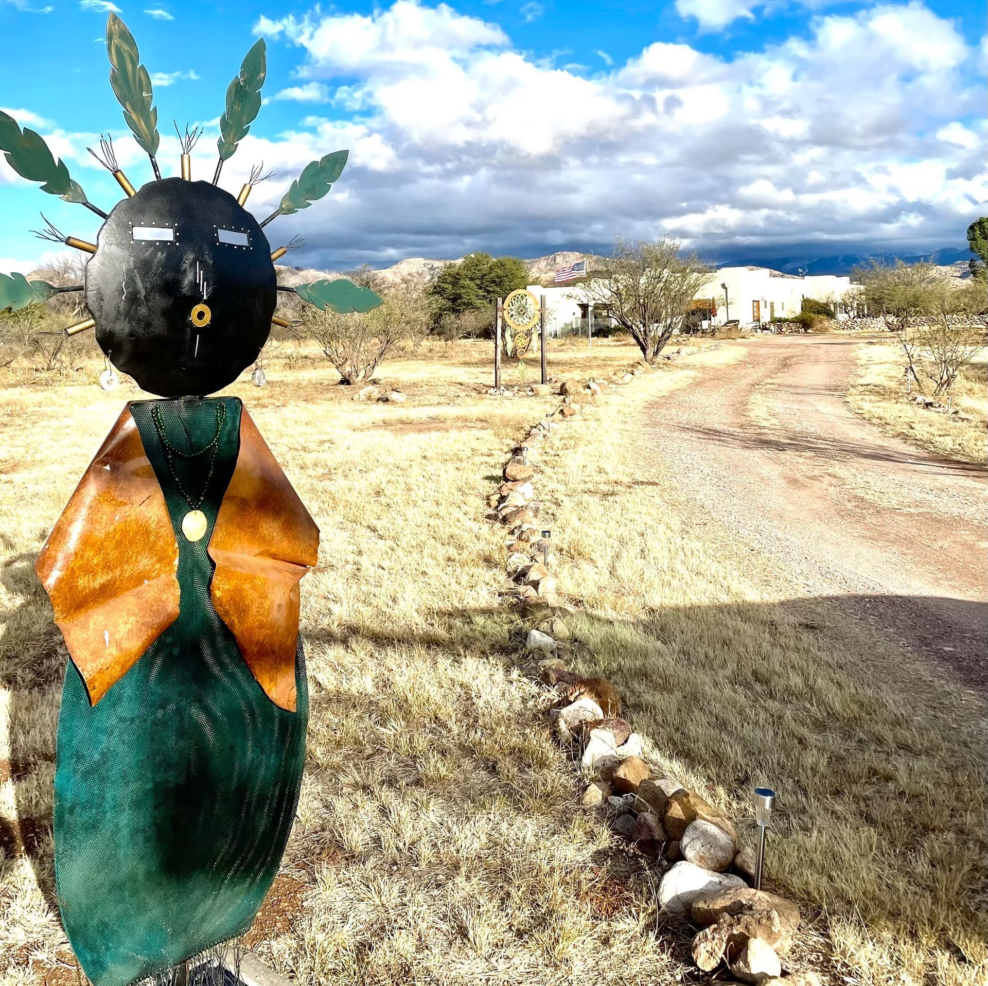 Metal figure with black head and green body beside a dirt road in a dry, rural landscape under a cloudy sky