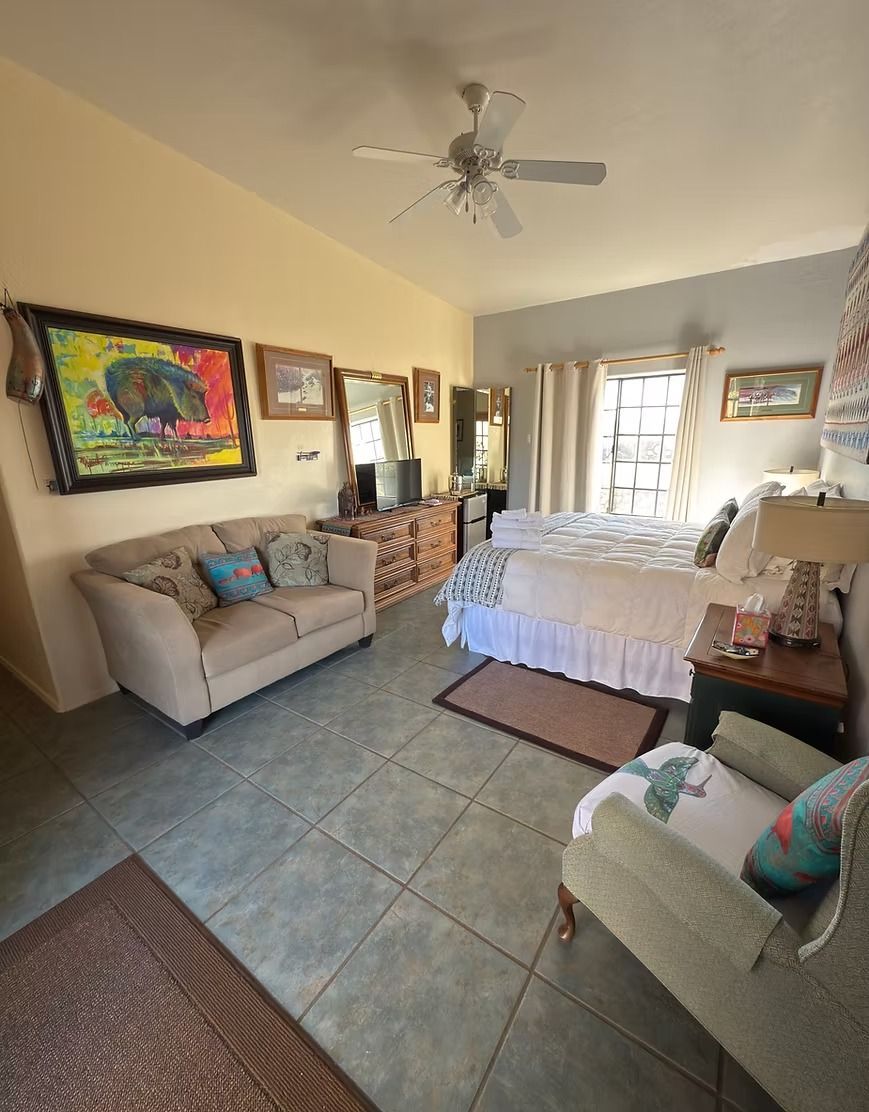 Bright bedroom with two beige chairs, a bed, dresser, tiled floor, and colorful wall art