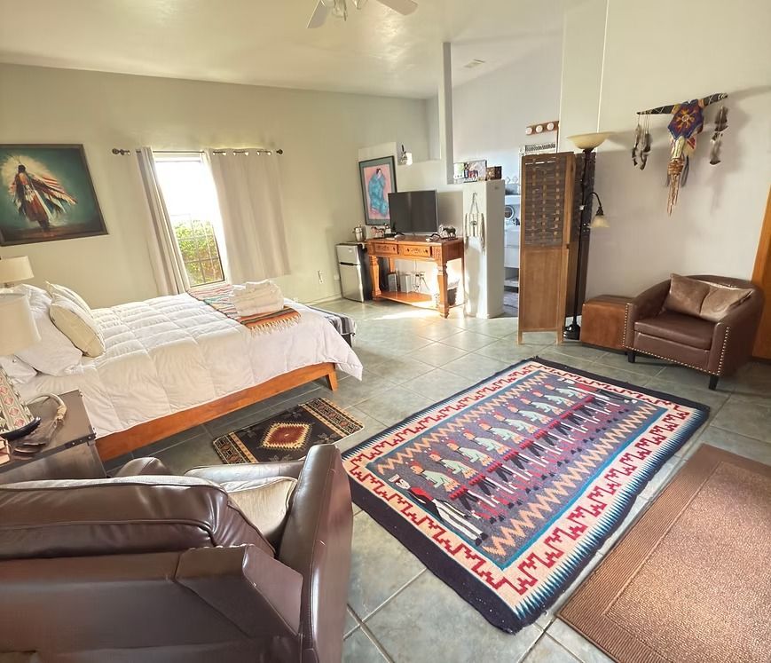Bright bedroom with a bed, leather chairs, colorful rug, and sunlight through the window.