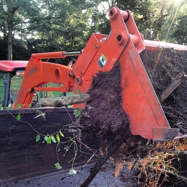 Orange excavator bucket clearing dark soil and roots beside a red truck in a wooded area