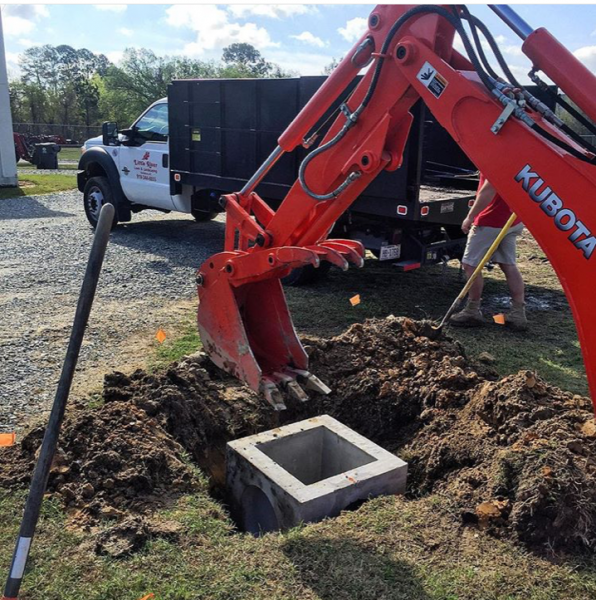 Orange Kubota excavator digging around a concrete utility box at a worksite, with a truck in the background