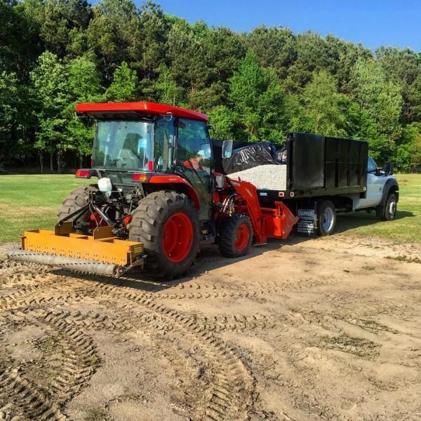 Red tractor grading dirt beside a black dump trailer and pickup truck on a sandy worksite