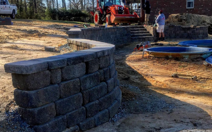 Curved stone retaining wall under construction beside a dirt path, with a red excavator and workers in the background