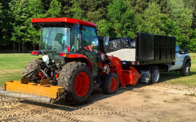 Red tractor with front attachment pulling a black dump trailer on a dirt path by green trees.