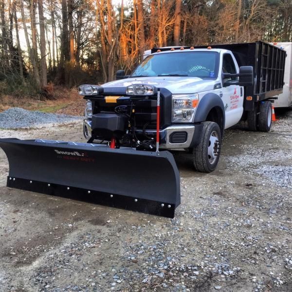 White utility truck with front plow parked on gravel in a wooded area at dusk