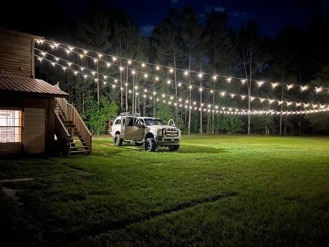 White SUV parked on a lit lawn beside a cabin under string lights at night