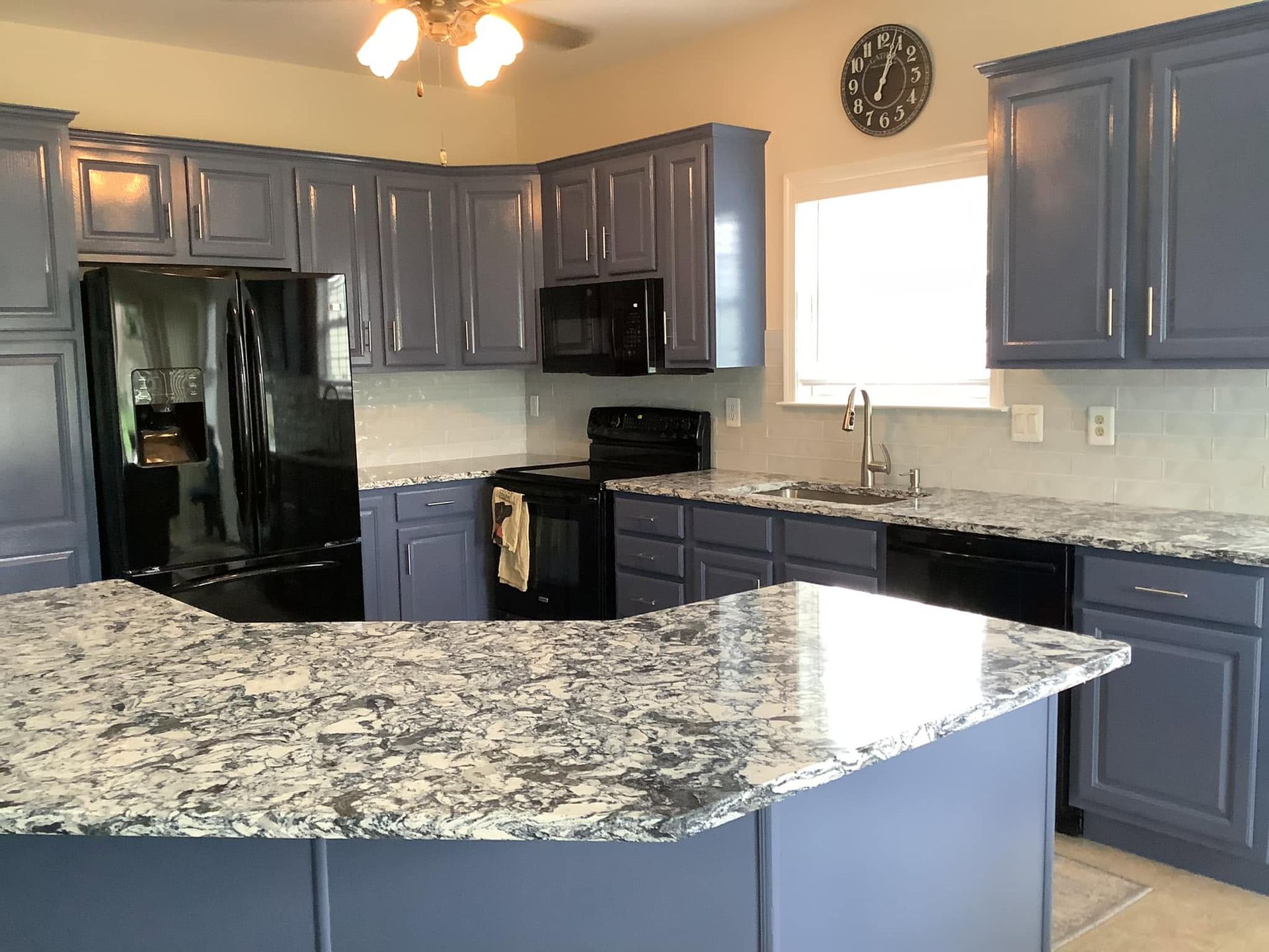 Modern kitchen with blue-gray cabinets, black appliances, and a large speckled granite island