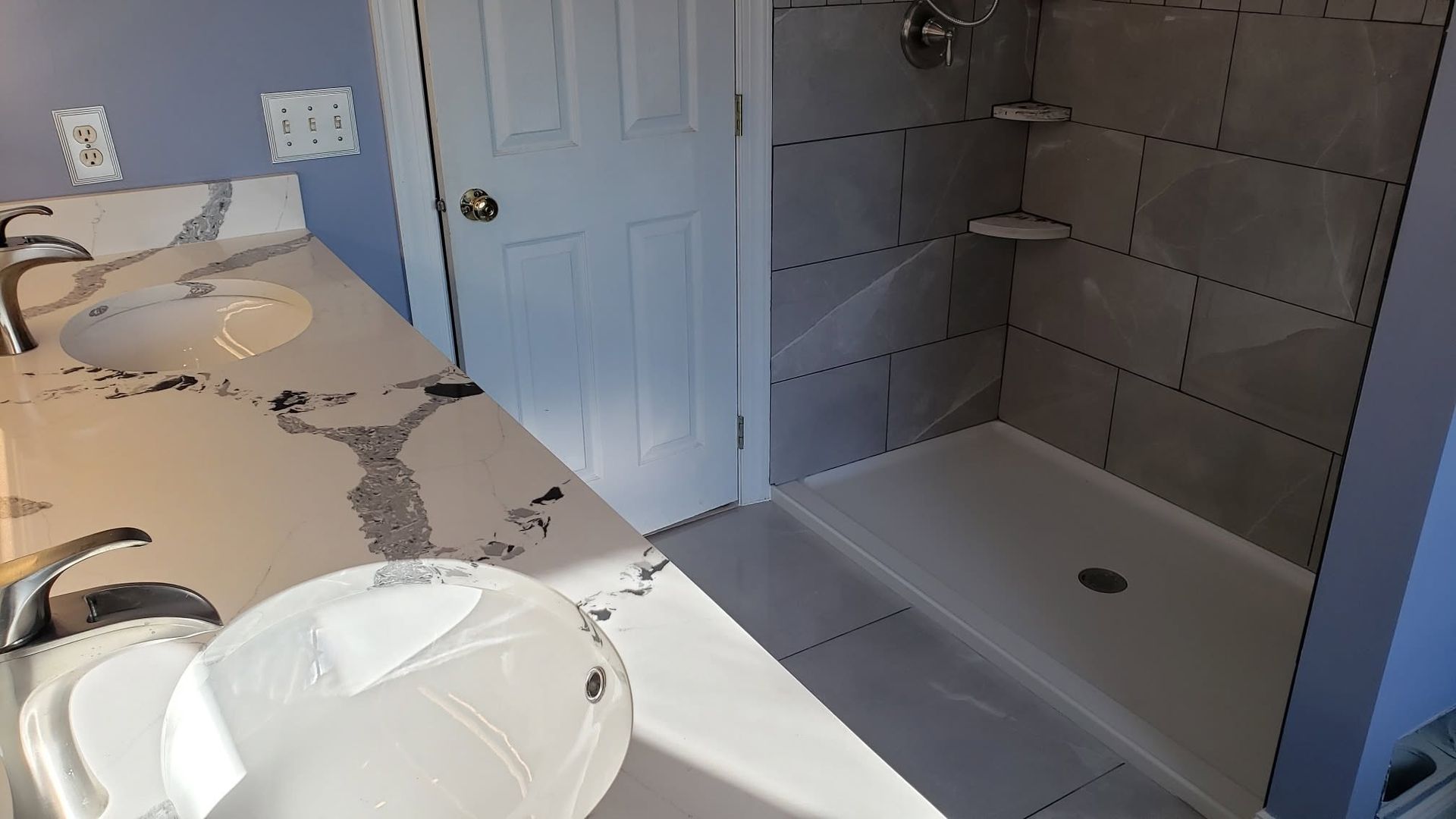 Bathroom vanity with two sinks beside a tiled walk-in shower and white door