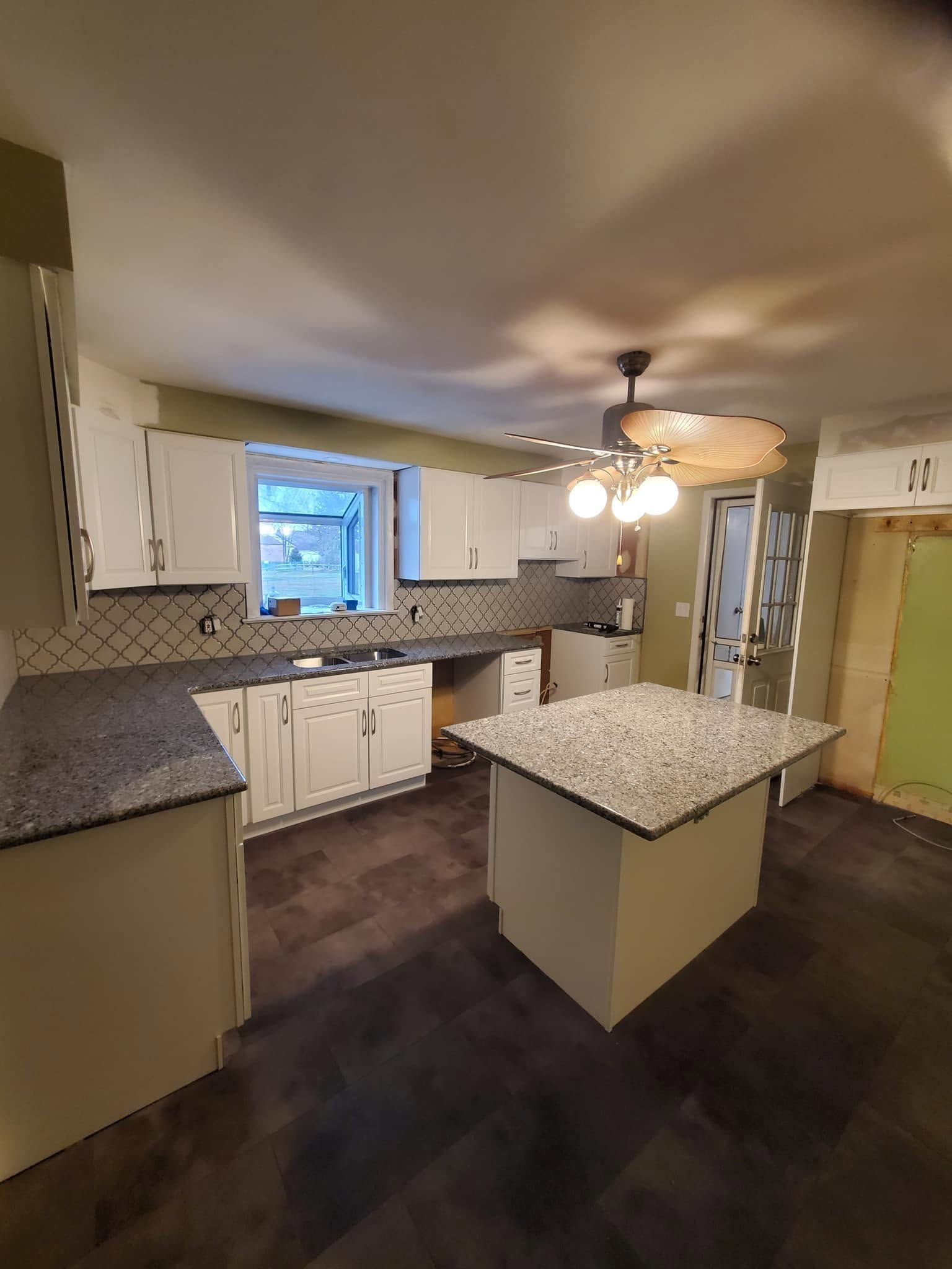 Empty kitchen with white cabinets, granite countertops, island, and ceiling fan under warm lights.
