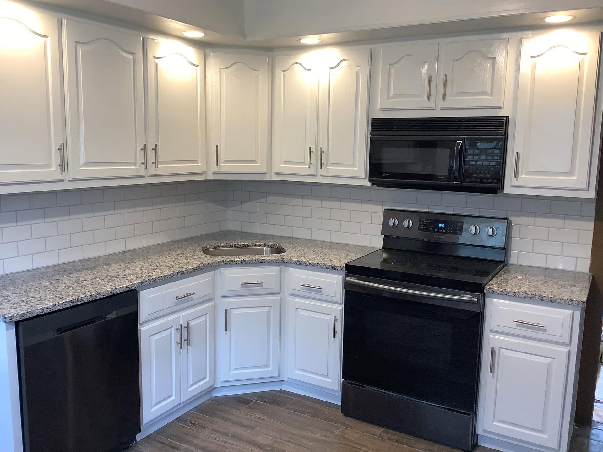 White kitchen with granite countertops, white cabinets, subway tile backsplash, black appliances, and dark floor.
