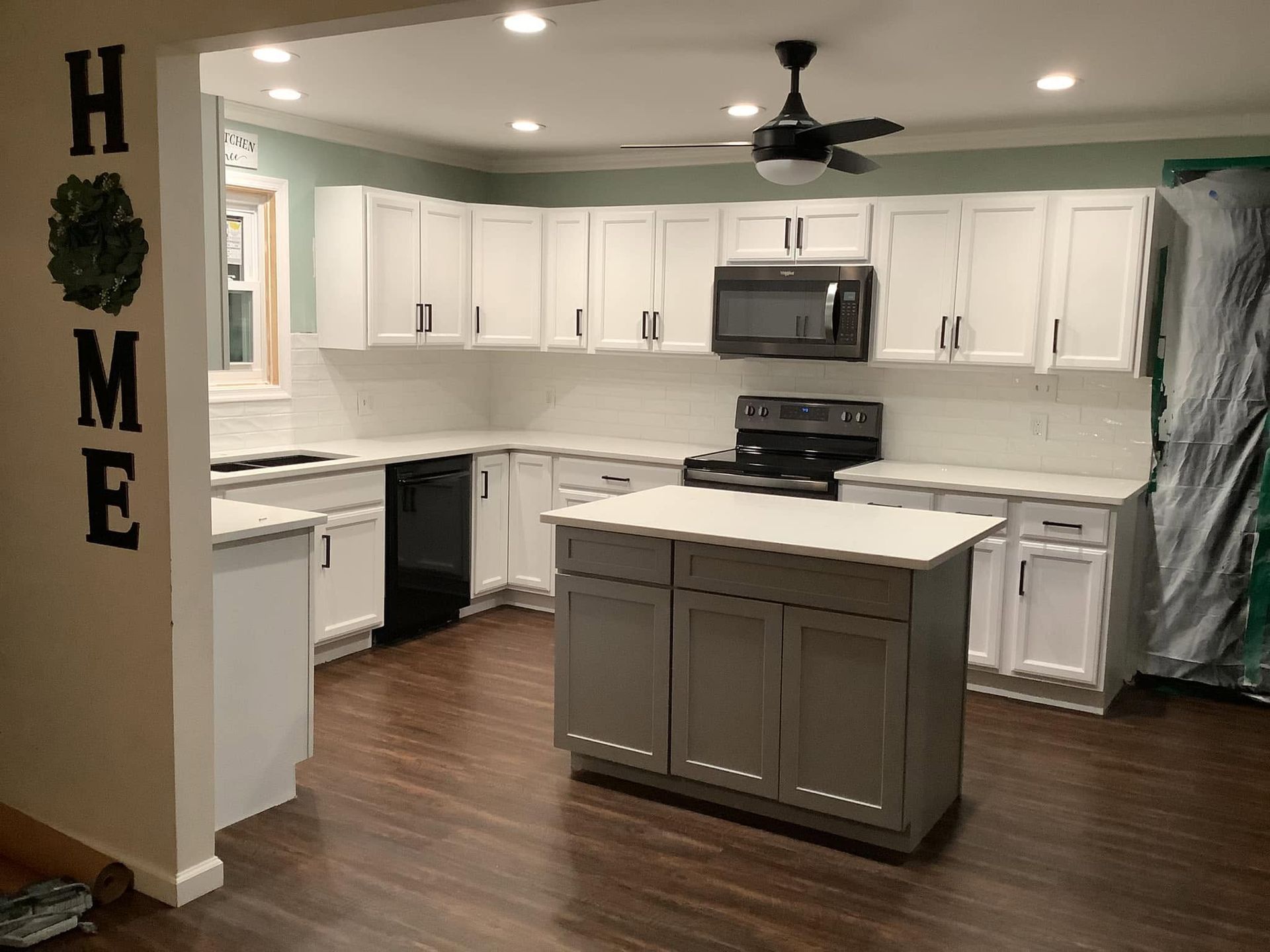 Bright kitchen with white cabinets, stainless appliances, gray island, and “HOME” wall decor on the left.