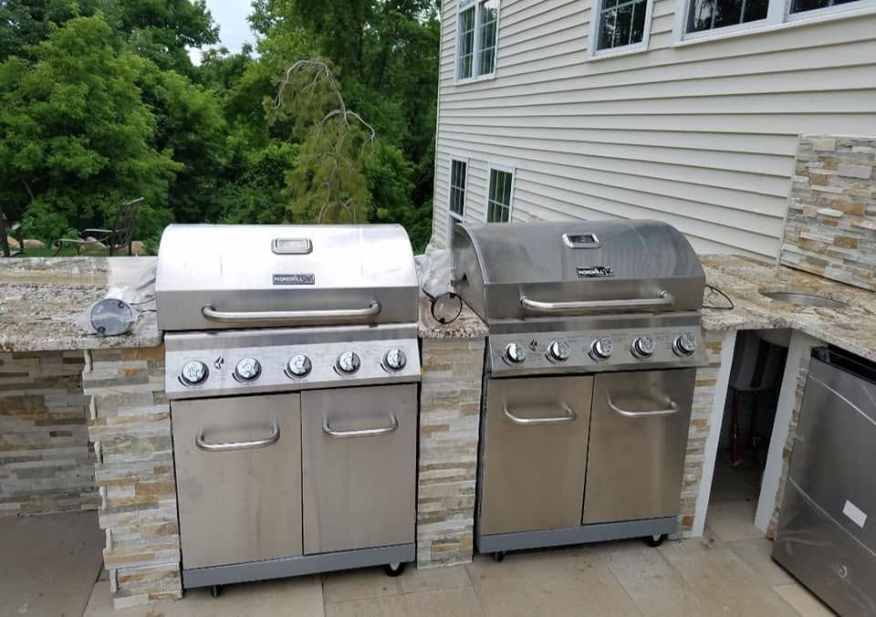 Two stainless steel gas grills on an outdoor patio beside a stone wall and house siding