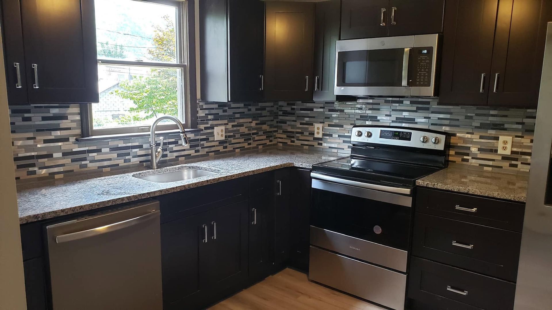 Modern kitchen with dark cabinets, stainless appliances, mosaic backsplash, and a window over the sink