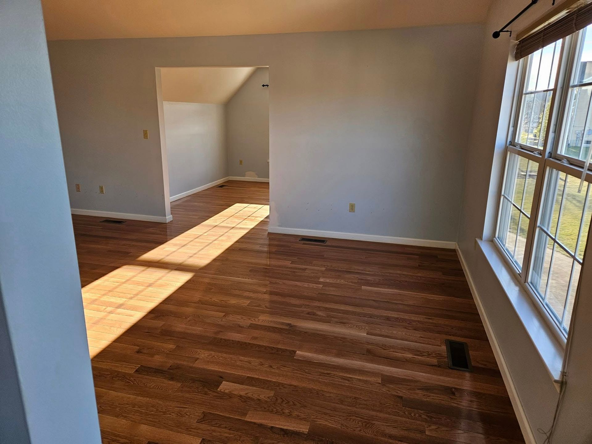 Empty sunlit living room with hardwood floors, beige walls, and large windows on the right.