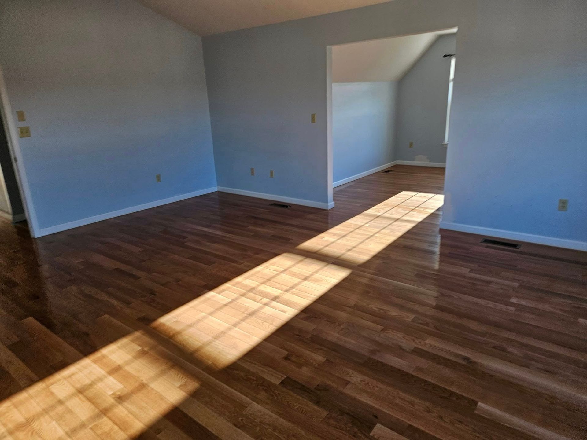 Empty room with hardwood floors, white walls, and sunlight streaming through windows.