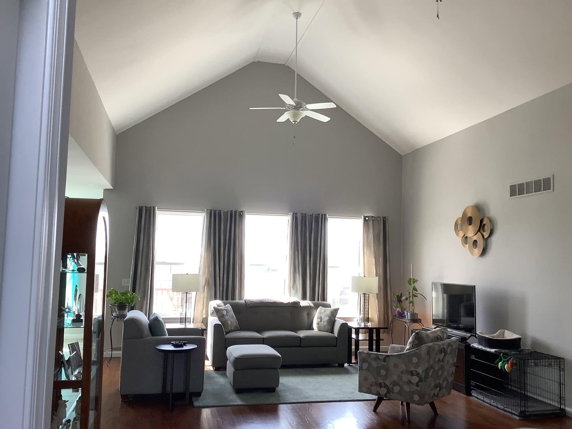 Living room with vaulted ceiling, gray sofas, large windows, ceiling fan, and TV on the right.