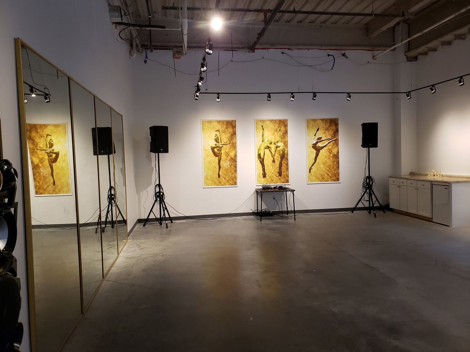 Art gallery with three gold paintings, speakers on stands, track lighting, and a mirrored wall.