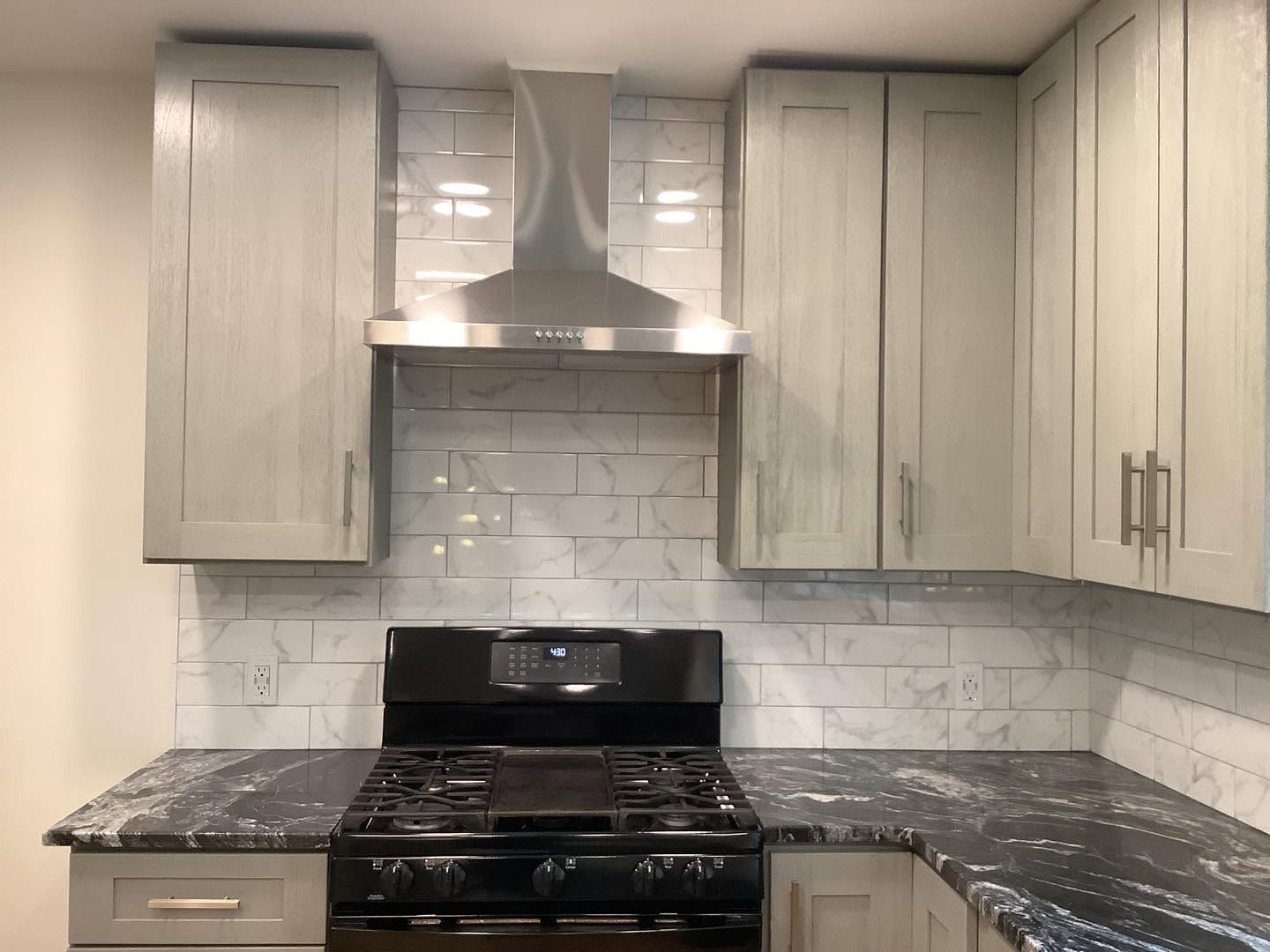 Modern kitchen with light gray cabinets, marble-pattern backsplash, black stove, and stainless range hood