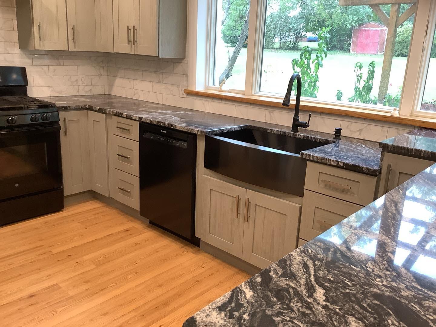 Modern kitchen with light wood cabinets, black countertops, stainless appliances, and large window over the sink