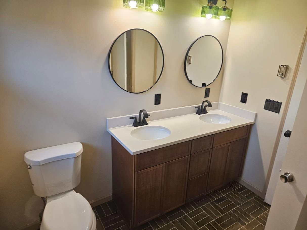 Small bathroom with toilet, double vanity sink, round mirrors, and wood cabinets on dark tile floor.