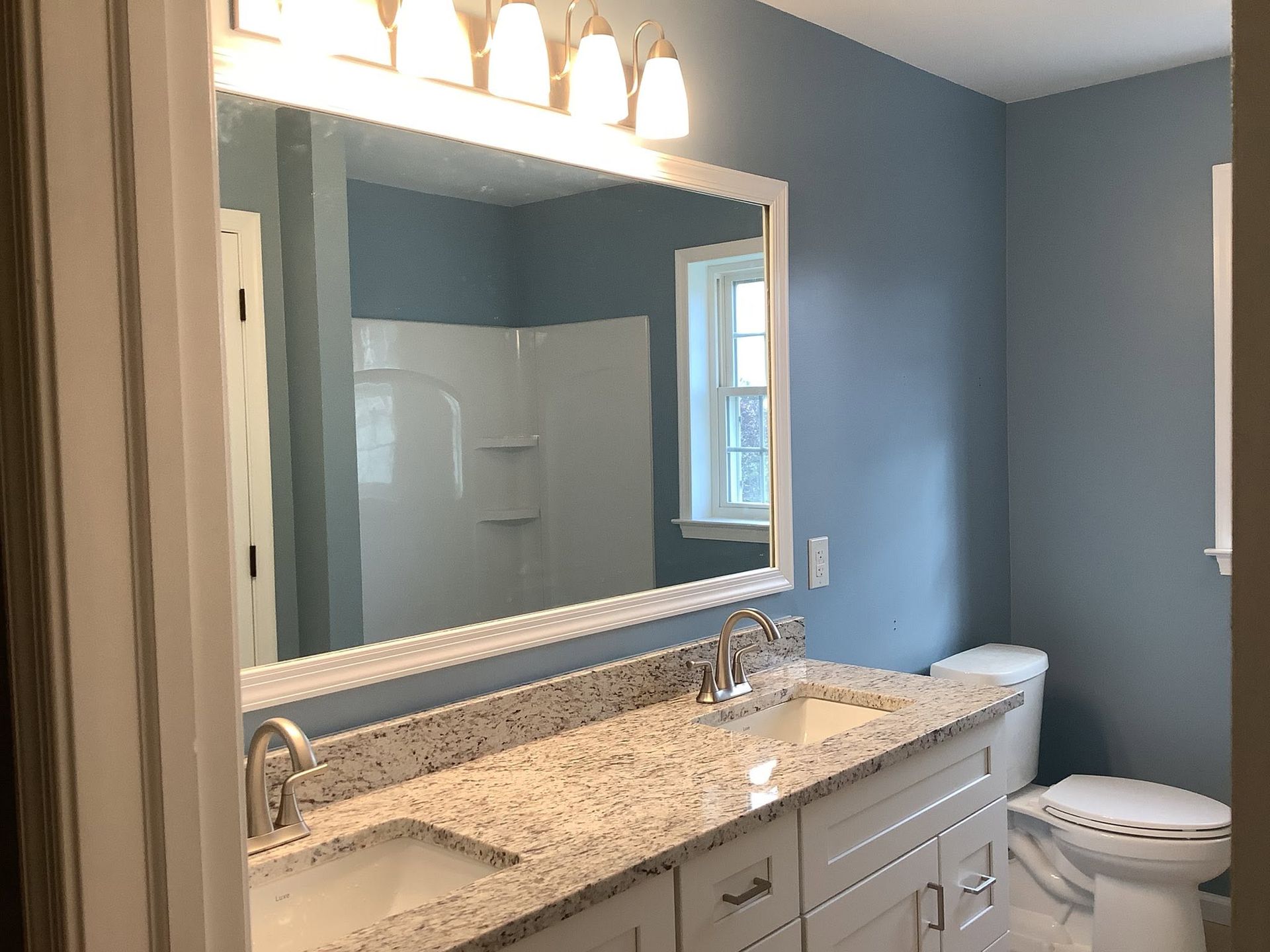 Bathroom with blue walls, large mirror, granite vanity, and a toilet near a window.